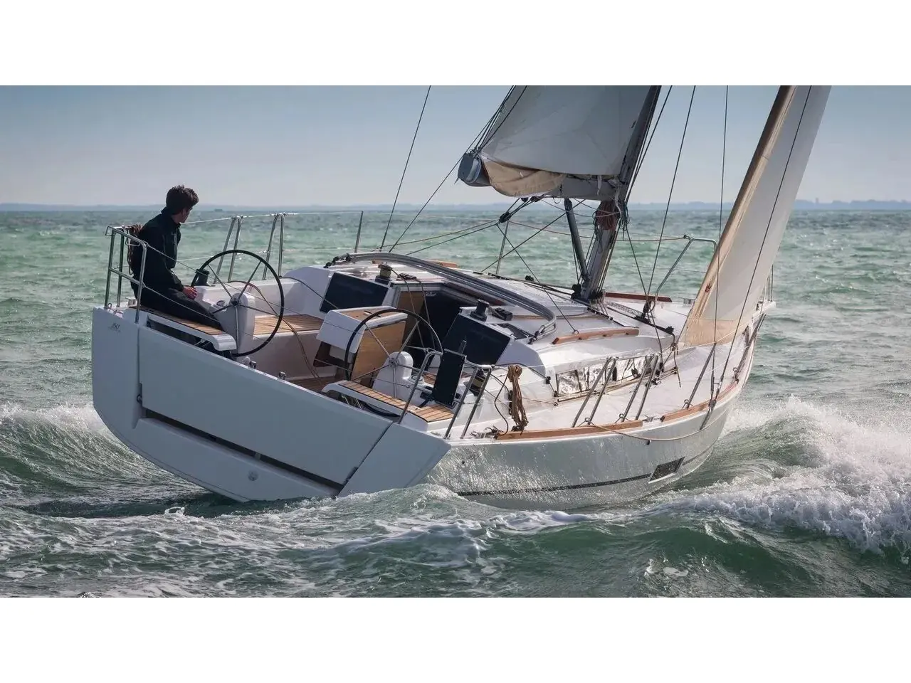 Un voilier monocoque blanc gîte sous voiles, avec une personne assise à la barre et des vagues qui déferlent le long de la coque.