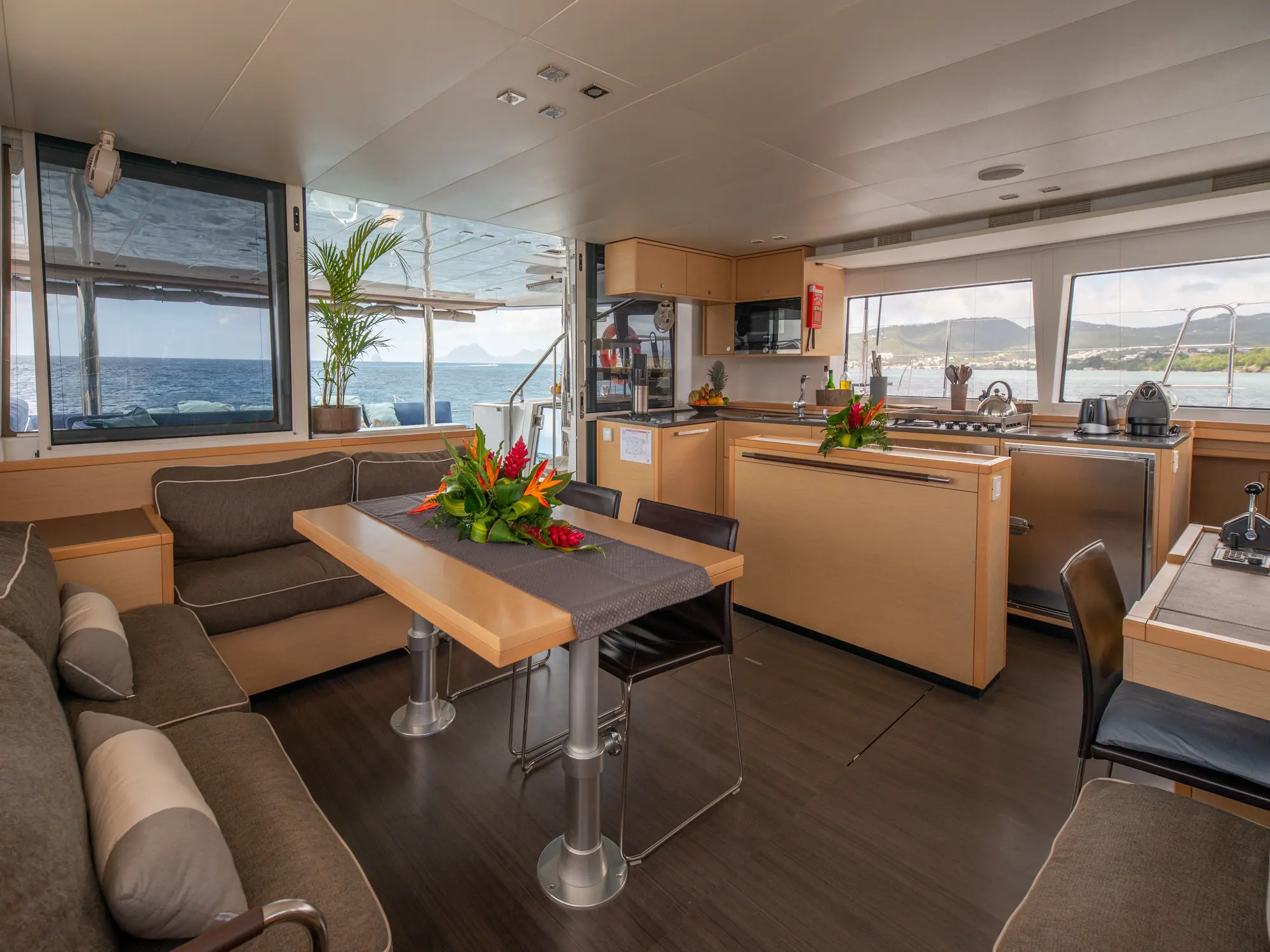 Carré de bateau avec banquette enveloppante, table et chaises, cuisine attenante, grandes fenêtres et ouverture vers un espace extérieur couvert.